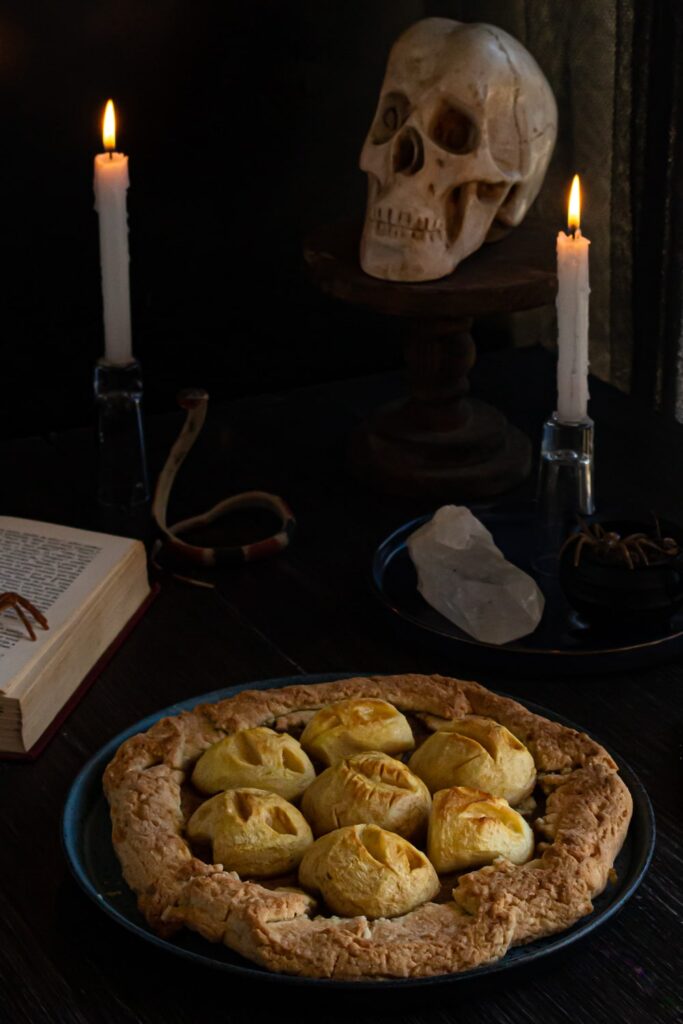 foto de uma torta com maçãs esculpidas como carinhas no topo, atrás da torta tem um crânio de gesso, velas acesas e um cristal