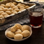 foto de um pratinho de biscoitos, com um deles mordido, e do lado uma xícara de chá escuro. Ao fundo um tabuleiro cheio de biscoitinhos