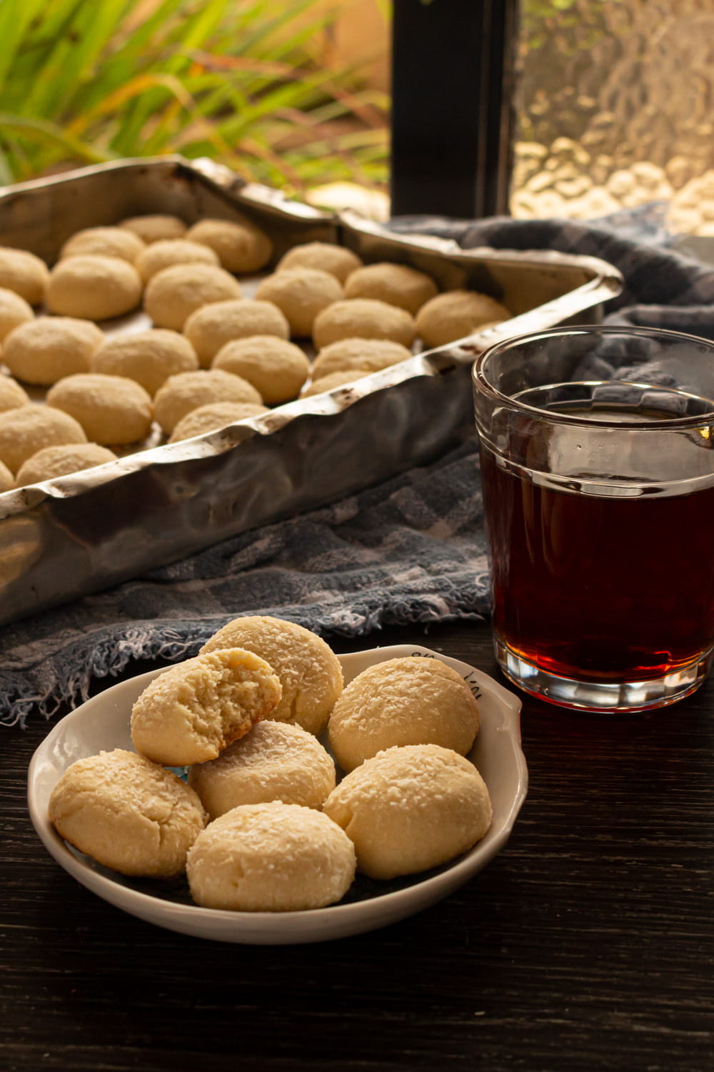 foto de uma mesa de madeira com um tabuleiro cheio de biscoitos, na frente dele um pratinho com mais biscoitos, um deles mordido, e do lado uma xícara de chá