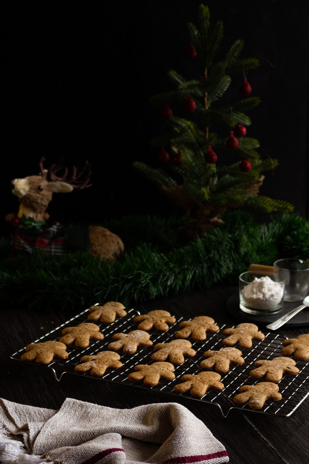 foto de uma mesa escura com uma bandeja de grade cheia de biscoitos de gengibre em formato de boneco. Atras dela um festão verde, um mini pinheiro com bolinhas vermelhas e um cervo de palha deitado do lado dele