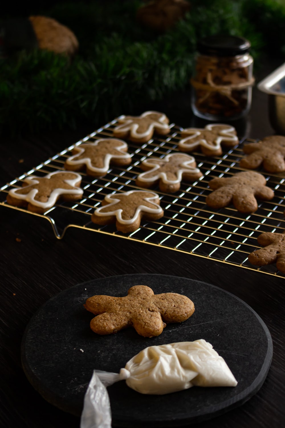 foto de um prato com um biscoito de gengibre e um saquinho com glacê branco. Atrás deles uma grade de metal com vários biscoitos já decorados sobre ela