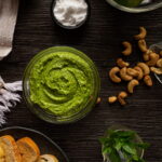 Tigela de pesto rodeada por um prato de torradas, uma tigelinha de sal, algumas folhas de manjericão e castanhas de caju