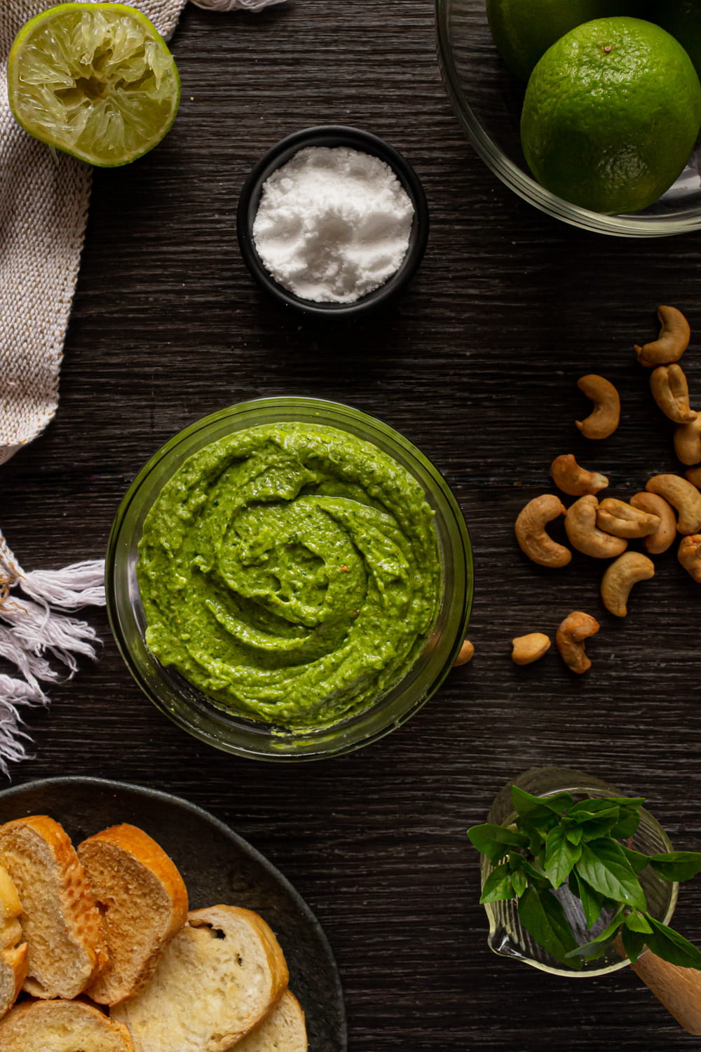vasilha com um molho liso verde, cercada por um prato com torradas, folhas de manjericão, limões, sal e castanhas de caju