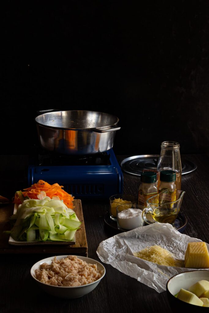mesa com ingredientes tigela de frango desfiado, queijo ralado, potes de temperos, e uma tábua com cenoura e abobrinha fatiadas