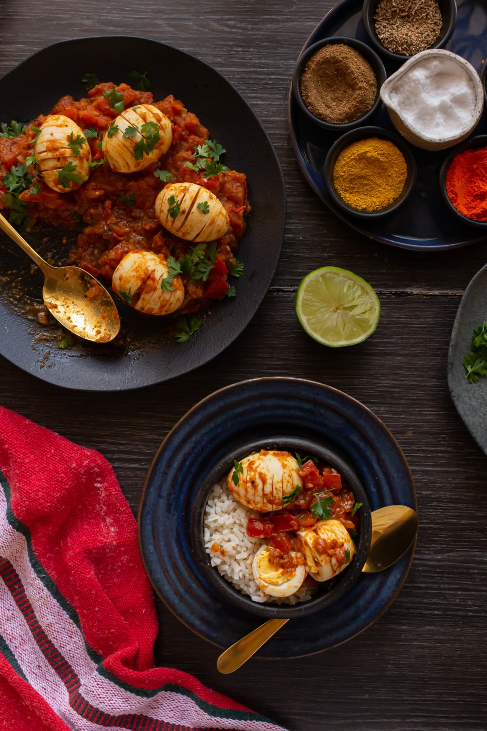 visão de cima da mesa de madeira com um prato escuro com o molho de tomate e os ovos cozidos por cima e um prato menor com uma tigela por cima, contendo arroz, um pouco do molho e dois ovos, um deles partido. Ao lado do prato tem maior outro menor, com vários potinhos com temperos em pó de cores diferentes, e ao lado dele um limão partido 
